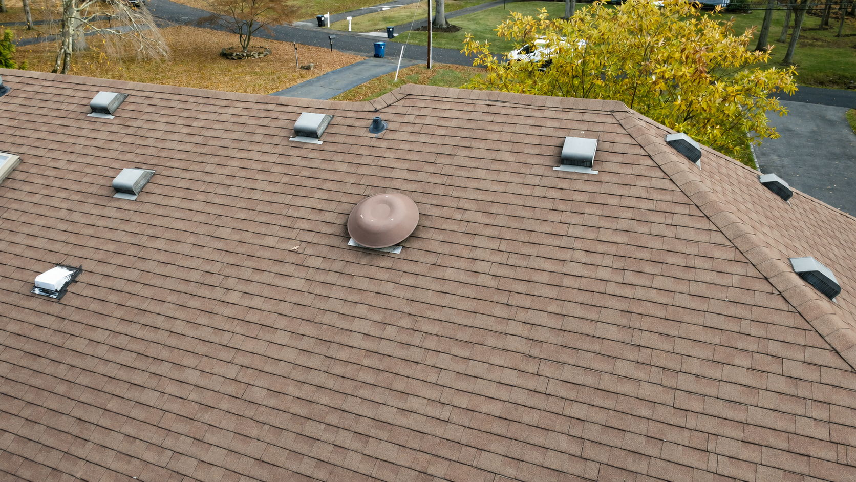 Rooftop with brown shingles, several skylight vents, and a round attic vent under a blue sky.