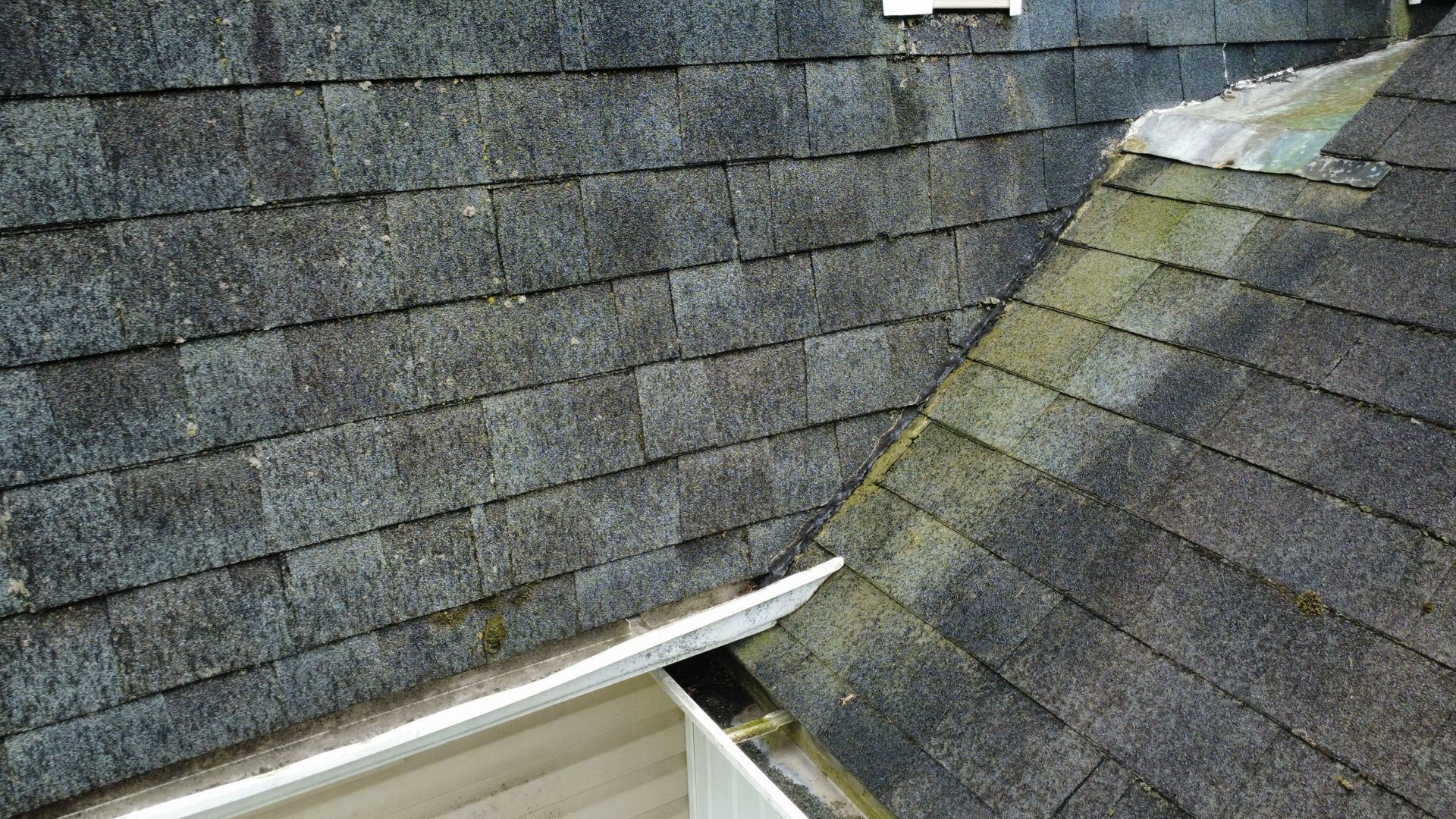 Weathered gray shingle roof with moss, showing a steep roof valley and gutter edge