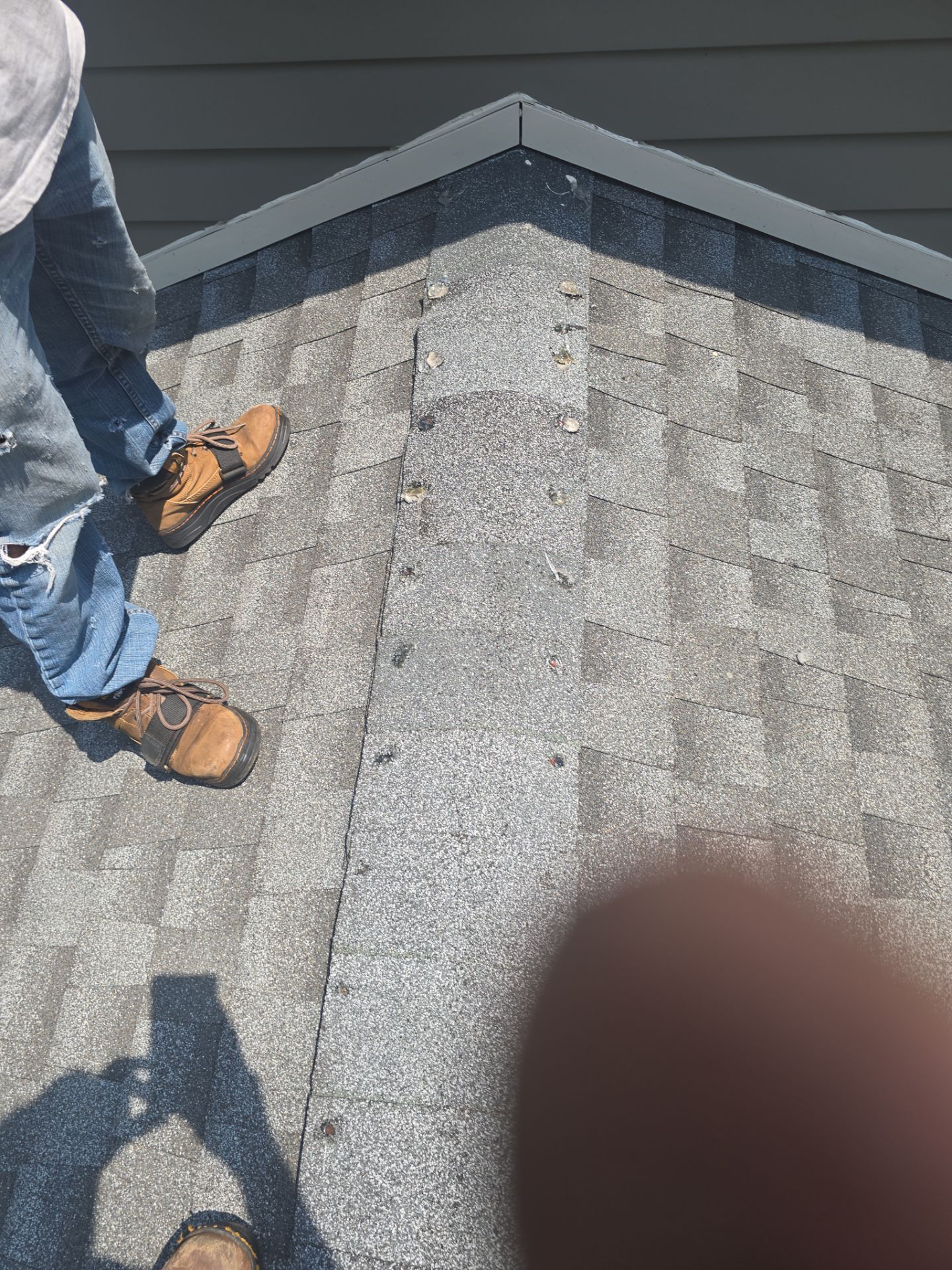 Person standing on a gray shingled roof near the edge, wearing jeans and brown shoes.
