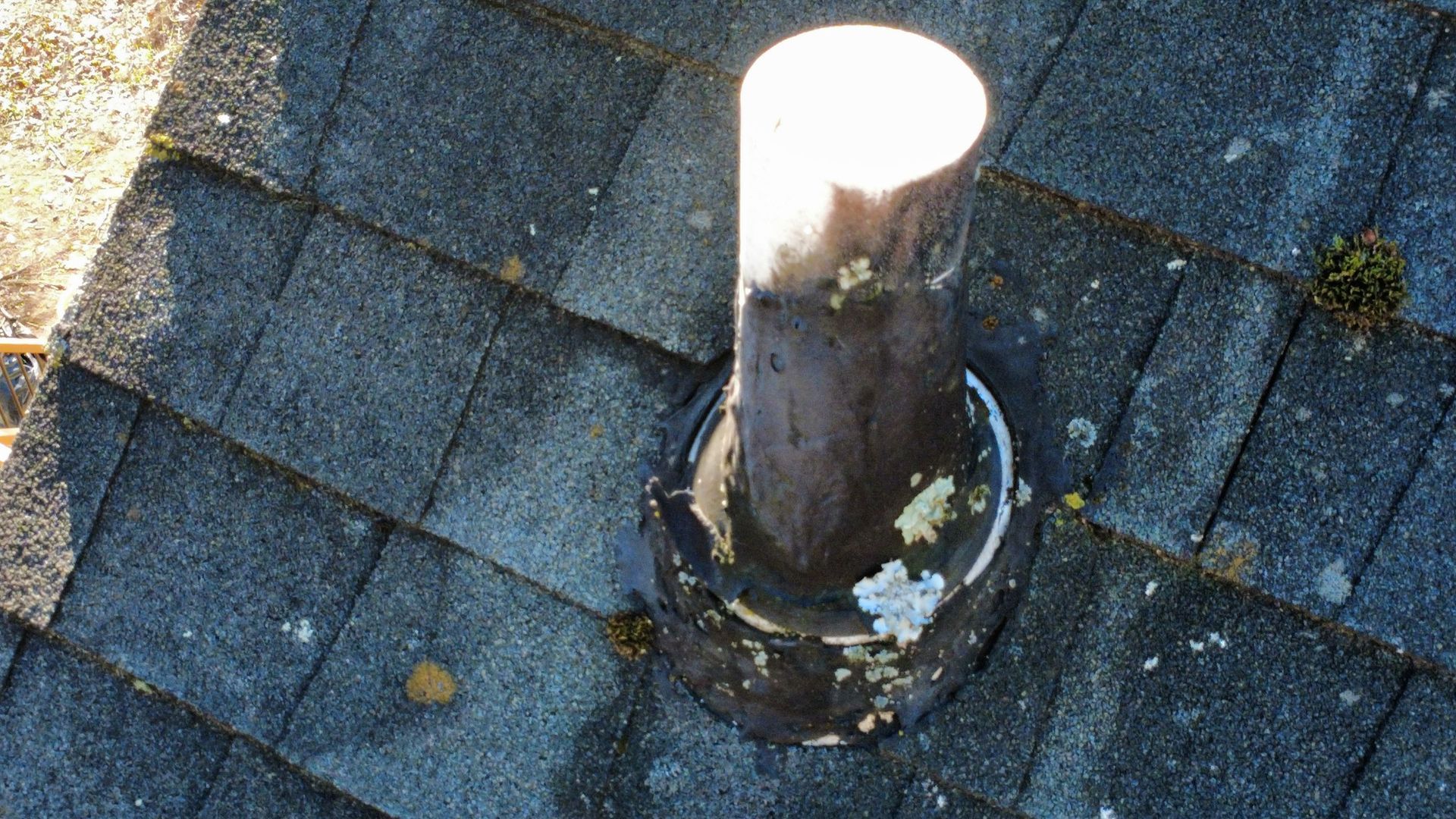 Roof vent pipe protruding through dark asphalt shingles, with white cap and weathered base.