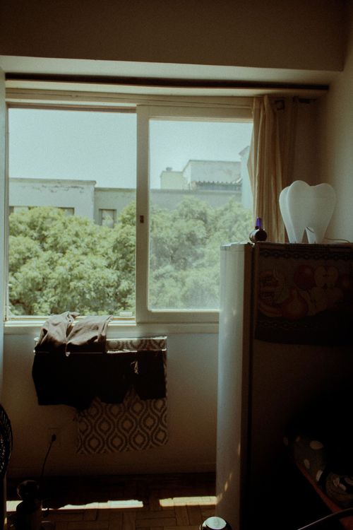 Dim bedroom with a large window, curtains, a dresser, and a suitcase near the bed.