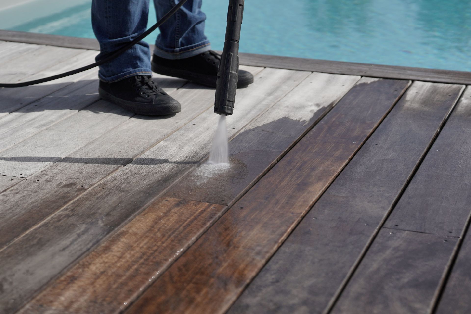 Person pressure-washing a wooden deck beside a pool