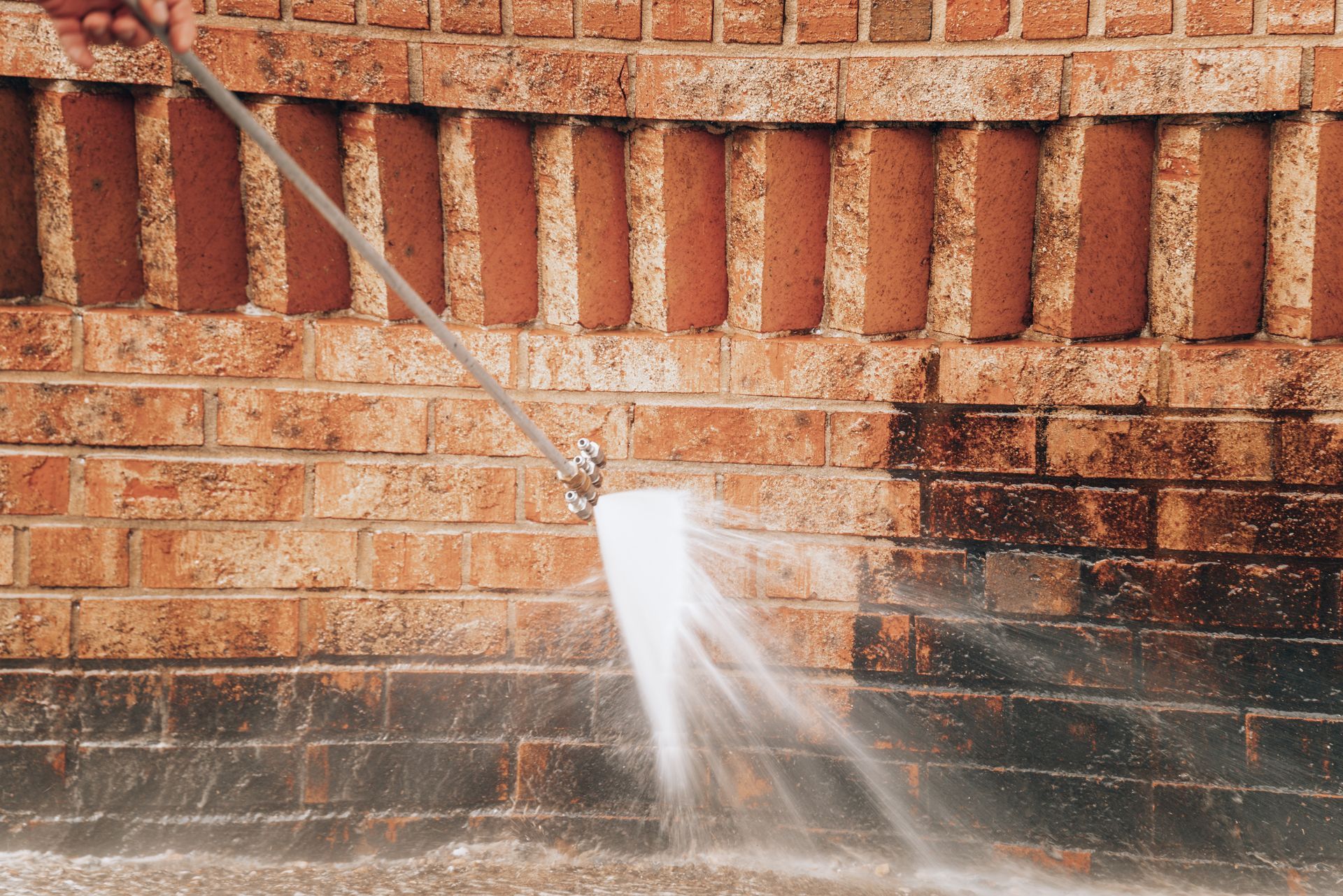 Person pressure-washing a red brick wall with water spray and splash on the ground