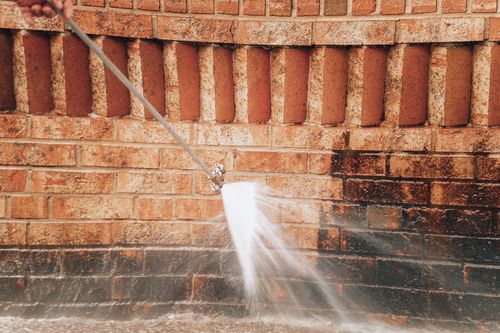 Person pressure-washing a red brick wall, spraying water across the lower bricks and pavement