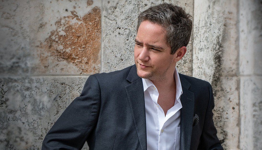 Man in blazer and open-collared shirt looking to the side, leaning against a stone wall.