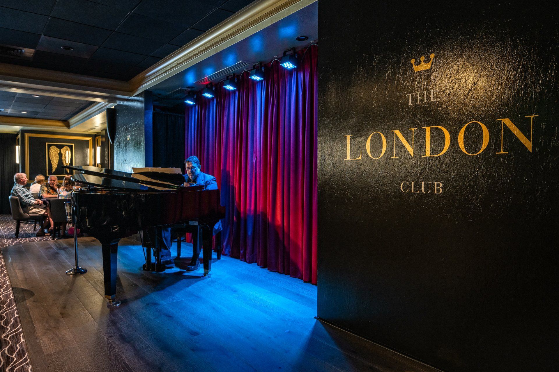 A pianist plays in front of red curtains at the London Bar. Dimly lit bar with patrons and a grand piano.
