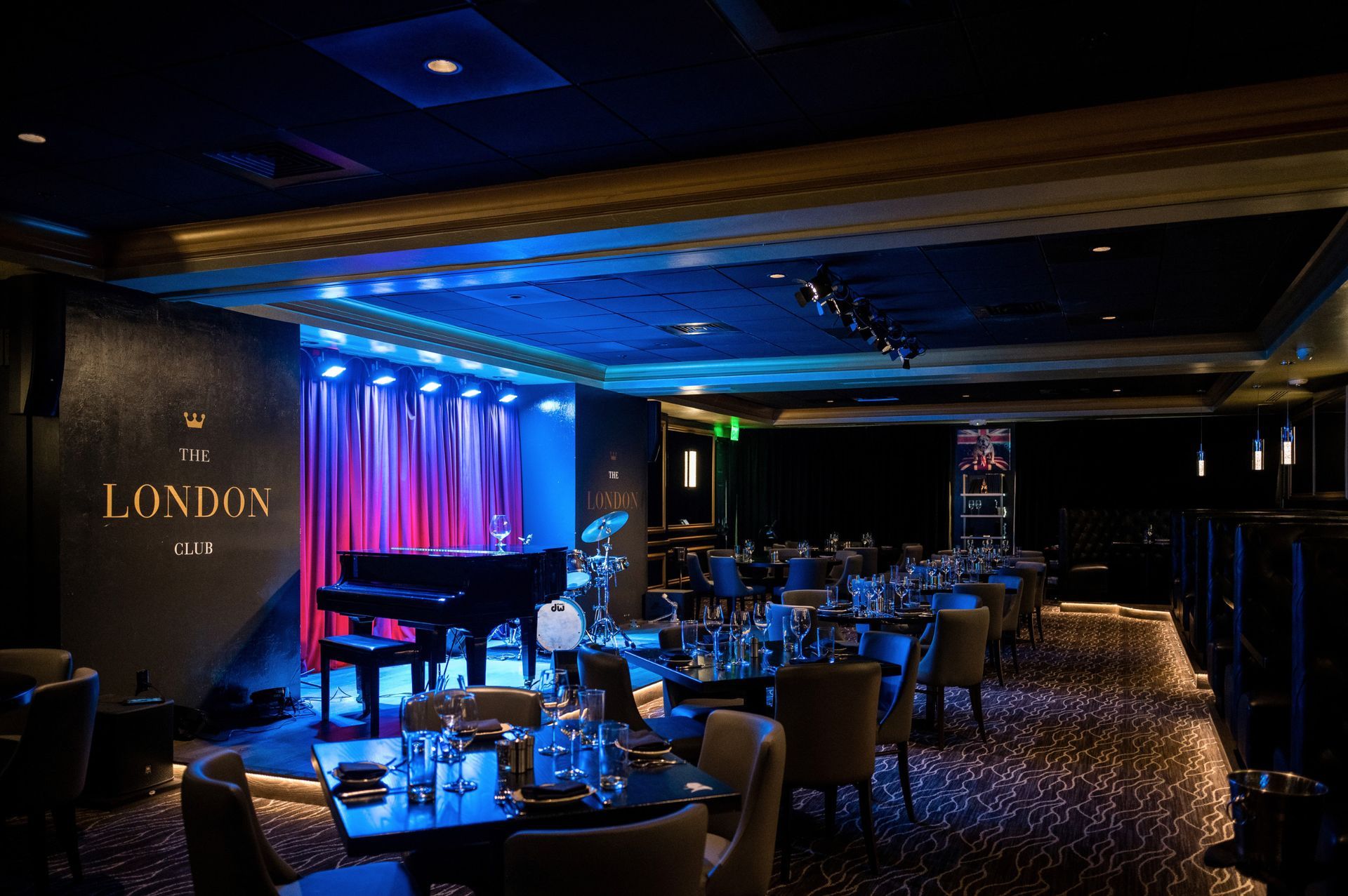 A dimly lit jazz club with a stage, piano, tables, and chairs. Blue and purple lighting creates ambiance.