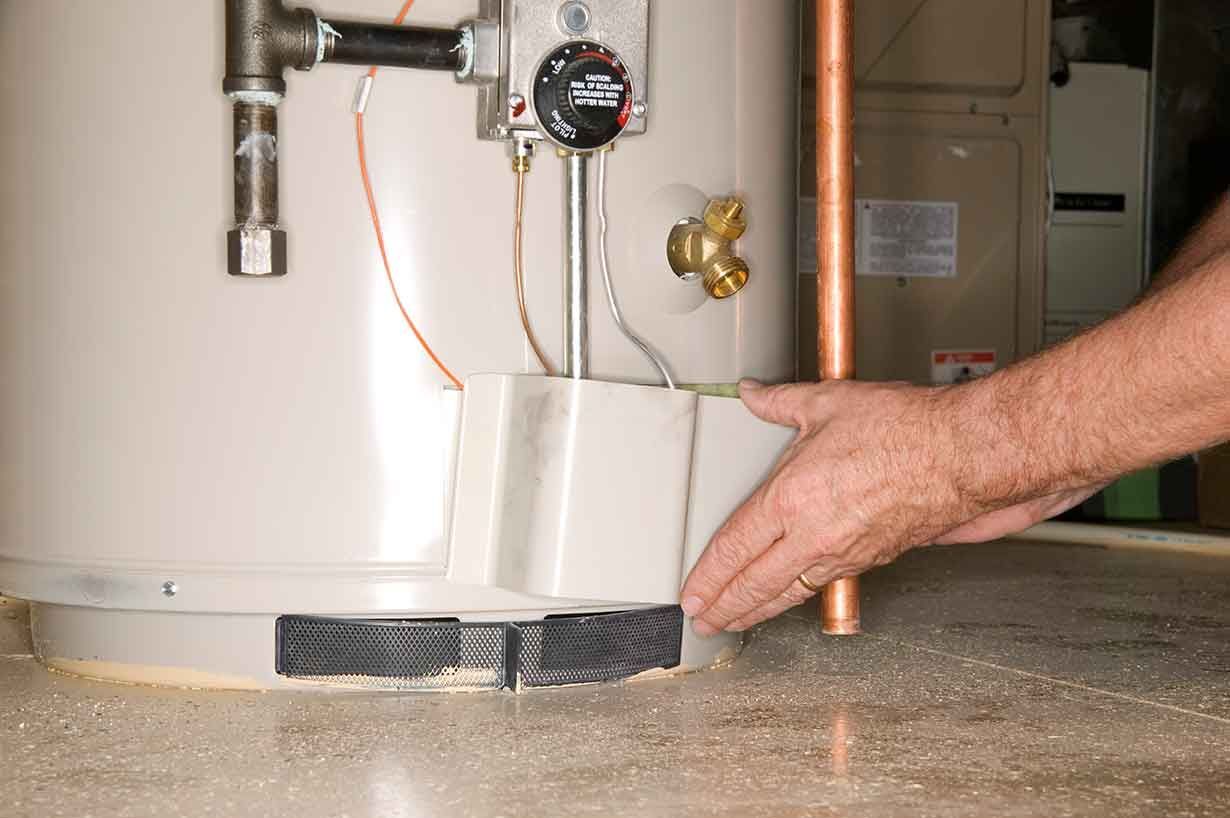Hand positioning a water heater on a basement floor near copper pipes and a pressure gauge