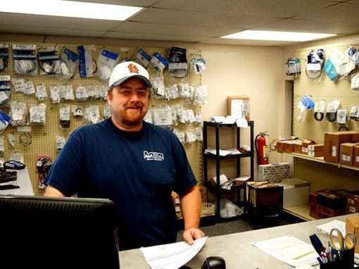 Man behind a counter in a small shop, with hanging merchandise and boxes in the background.