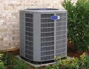 Gray outdoor air conditioner unit beside a brick wall and green shrubs