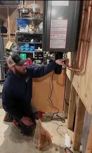 Technician kneels beside a boiler, inspecting copper pipes in a utility room.