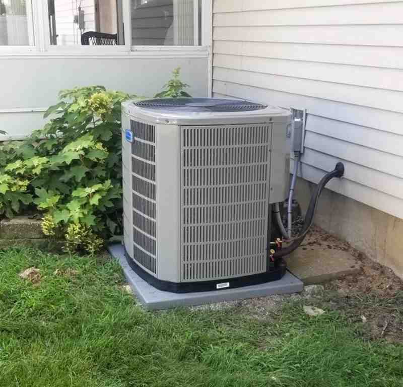 Outdoor central air conditioner beside a house, on a concrete pad near siding and shrubs