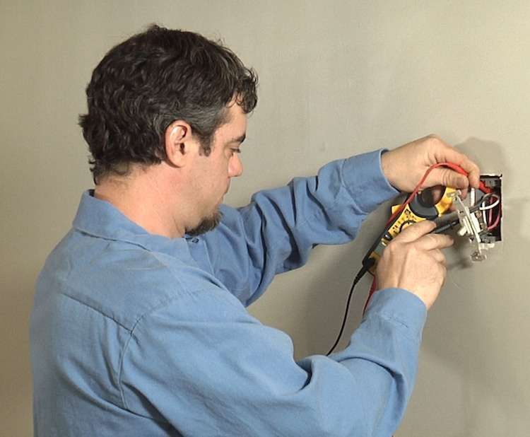 Man using a multimeter to test exposed electrical wiring in a wall outlet box