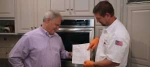 Two men in a kitchen review a recipe sheet, one pointing with orange gloves.