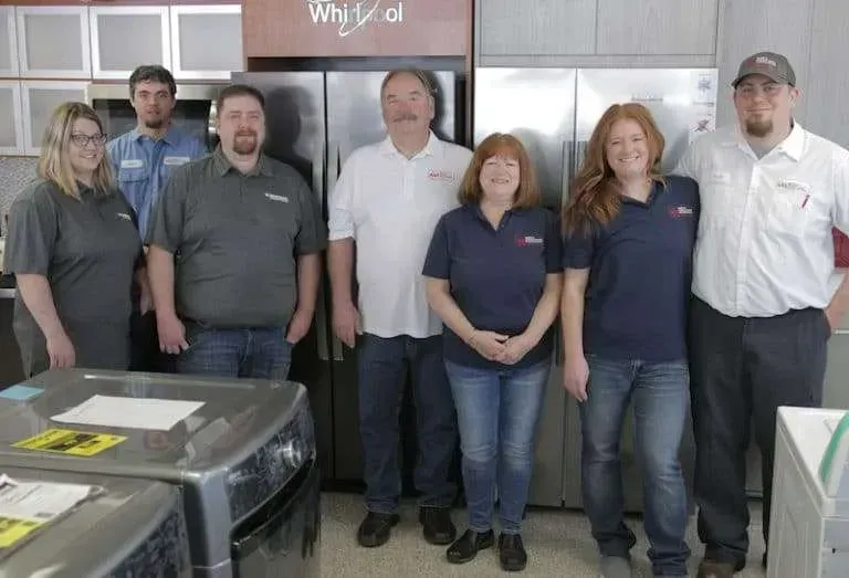 Group of six people standing in a kitchen, posing behind counters and cabinets.