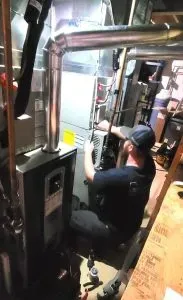 Worker in black shirt and cap crouches inside a metal machinery compartment, inspecting equipment.