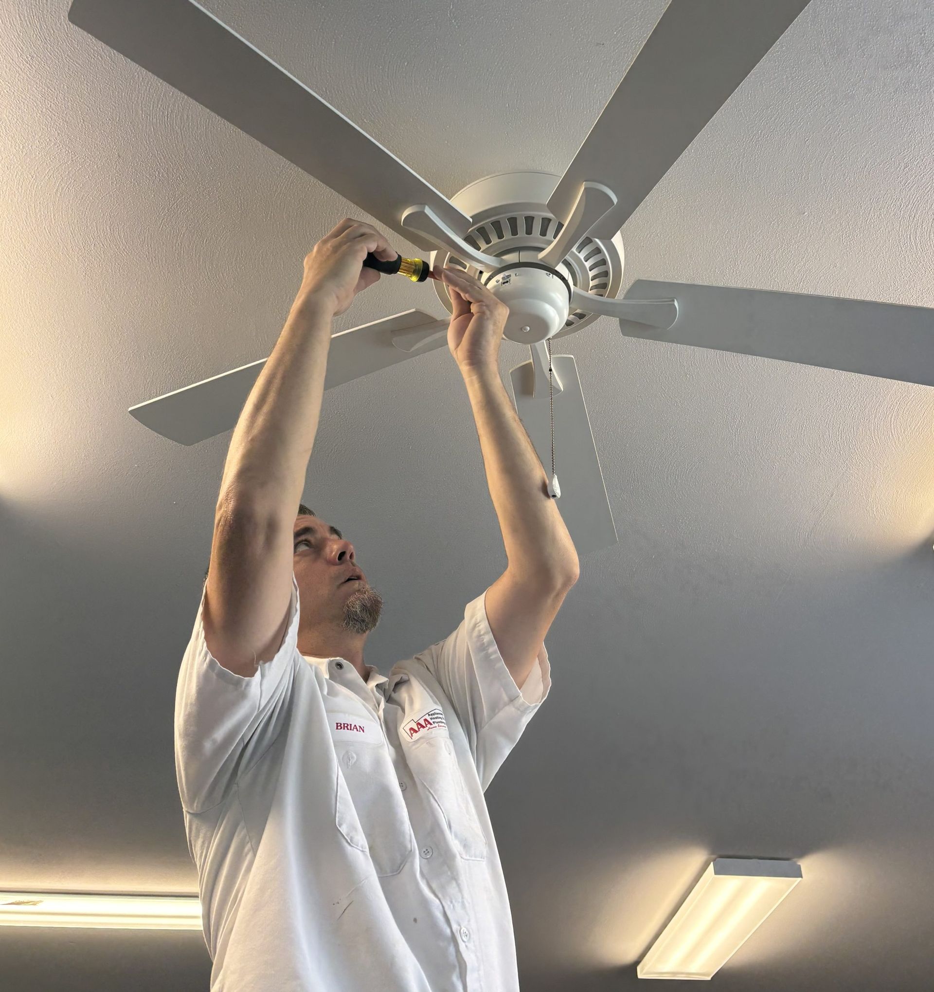 Technician adjusting a ceiling fan with a screwdriver indoors underneath fluorescent lights