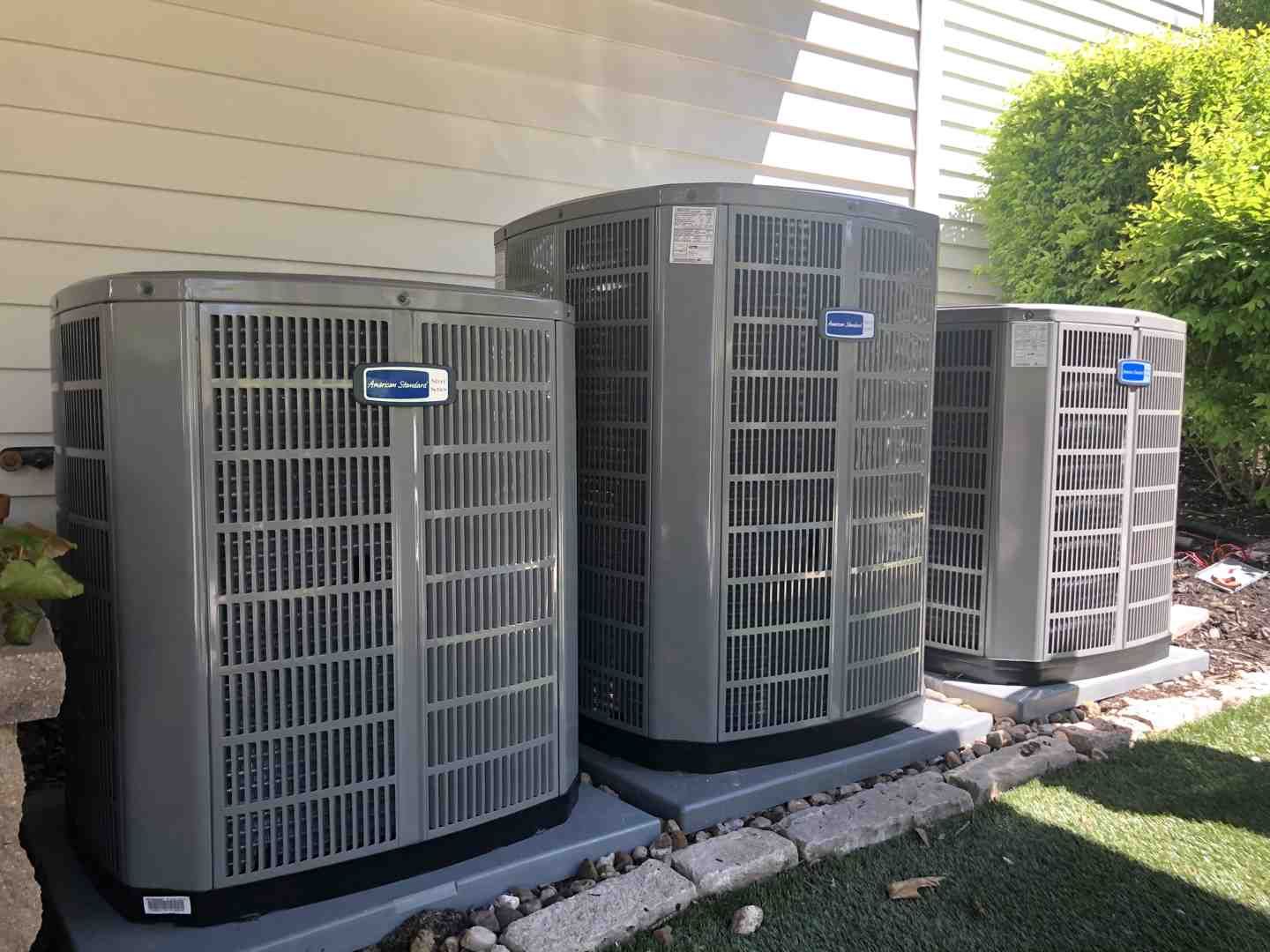 Three outdoor HVAC condenser units beside a house on gravel, with grass and bushes nearby
