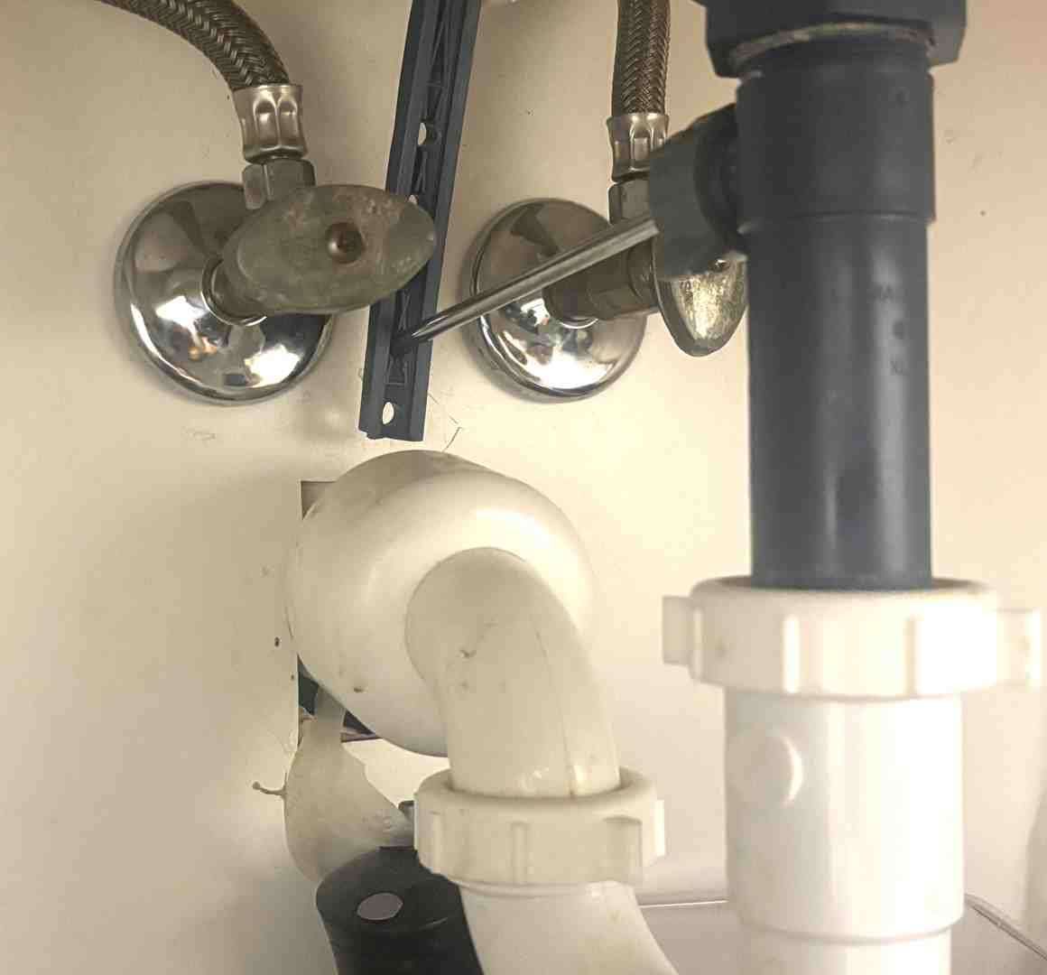 Under-sink plumbing with chrome water valves, braided hoses, and white PVC drain pipes