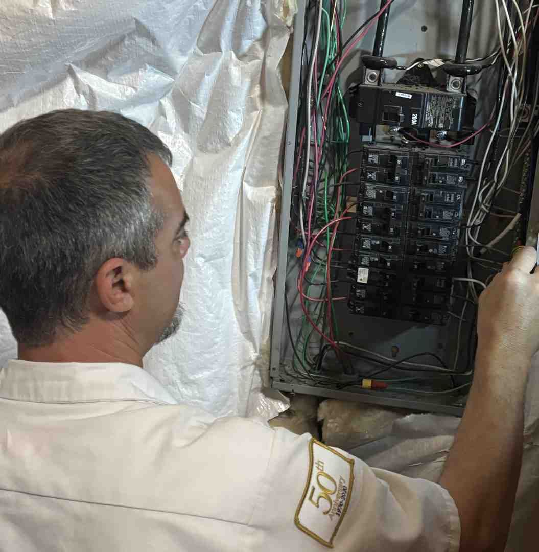 Technician inspecting an open electrical breaker panel with exposed wiring in a wall cavity
