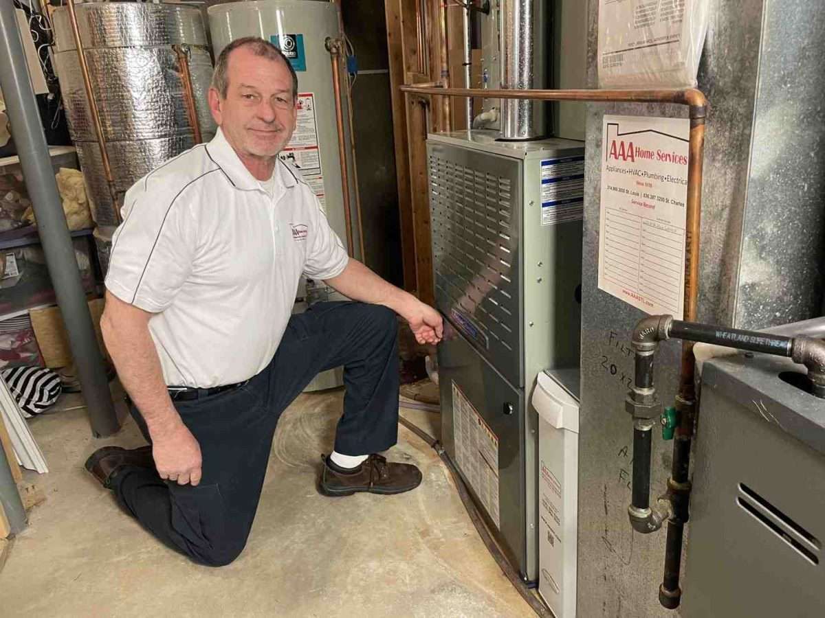 Technician kneeling beside a furnace and HVAC unit in a utility room, inspecting the equipment.