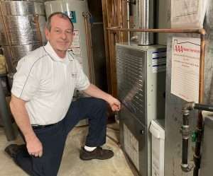 Man kneeling beside a furnace in a utility room, pointing at the equipment.