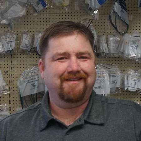 Smiling man in a gray polo shirt standing in front of a pegboard wall with hanging packages