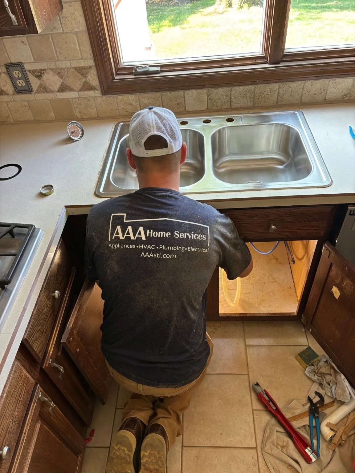 Person kneeling under kitchen sink, working on plumbing repair beside a double basin sink.