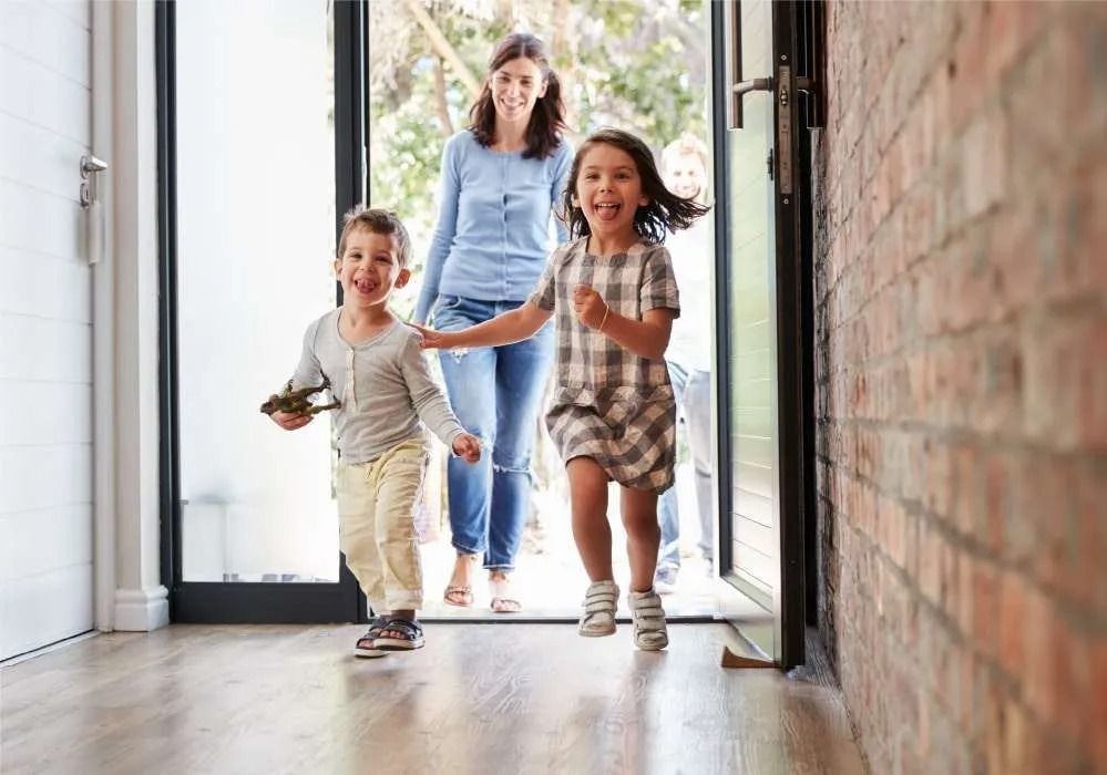 Two children running down a bright hallway while a woman follows behind, smiling