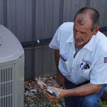 Technician crouches beside an outdoor HVAC unit, using a tool to inspect wiring by a metal building.
