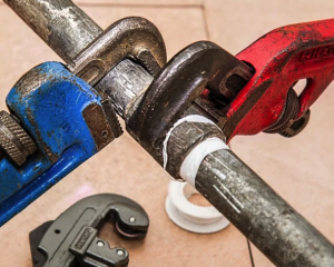 Three pipe wrenches gripping a metal pipe, with blue, black, and red handles on a tiled floor.