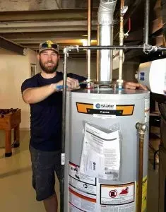 Man standing beside a large water heater in a basement, holding a tool near the unit