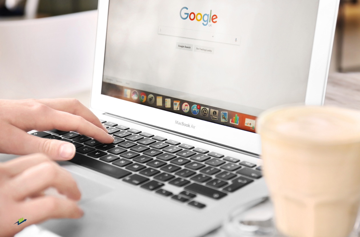 Person typing on a MacBook laptop, Google search bar open. Coffee cup sits nearby.