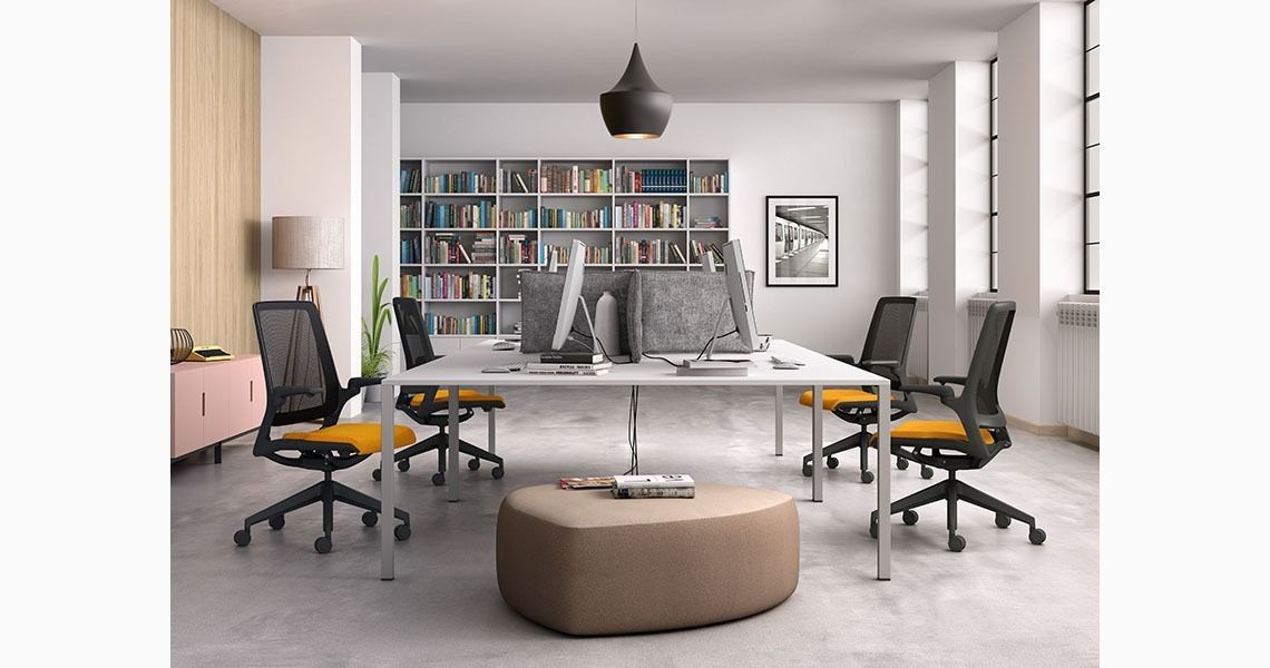 Un moderno spazio ufficio con una scrivania bianca per quattro persone, sedie ergonomiche con seduta gialla, un pouf centrale e librerie.