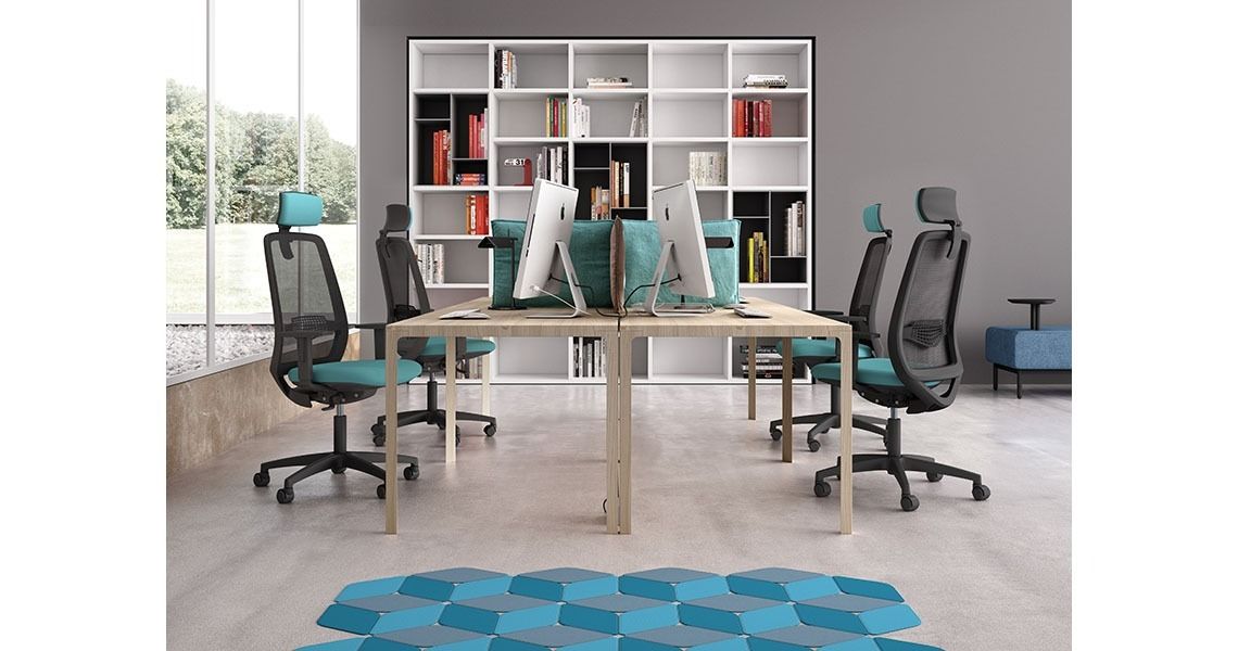 Un moderno spazio di lavoro in ufficio con quattro sedie ergonomiche in rete nera, due scrivanie contrapposte con computer e una grande libreria.