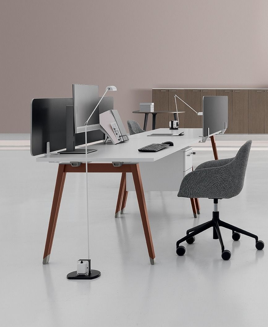 Postazione di lavoro moderna in ufficio con una lunga scrivania bianca condivisa, gambe in legno marrone, due monitor e una sedia con rotelle.