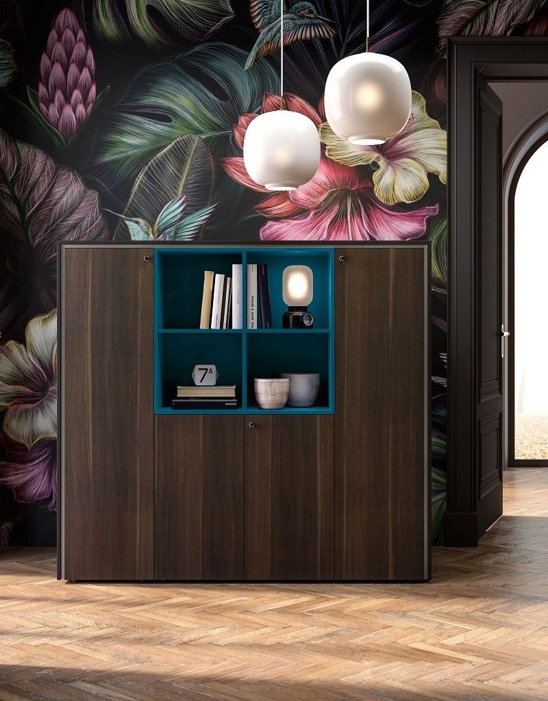In una stanza, un mobile contenitore in legno scuro con un ripiano interno a giorno color verde acqua fa da sfondo a una carta da parati floreale scura.
