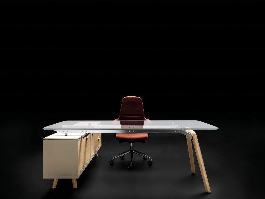Una moderna scrivania da ufficio con piano in vetro e gambe in legno chiaro è posizionata di fronte a una sedia da ufficio con rotelle color ruggine.