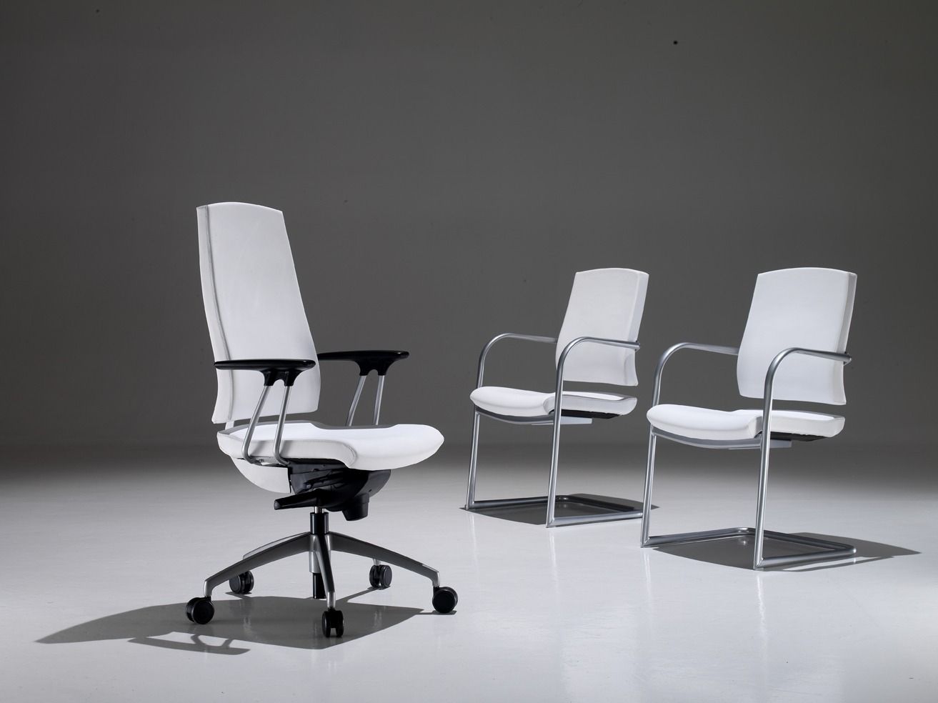 In un monolocale, una sedia da ufficio con ruote e due poltrone fisse, tutte con rivestimento bianco e struttura argentata.