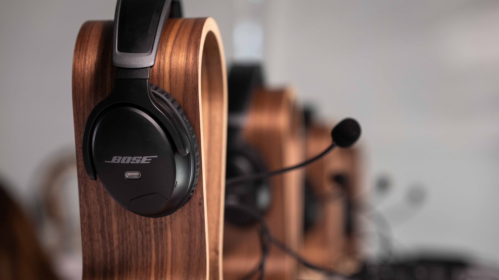 Black Bose headphones hanging on a wooden stand, with a blurred headset in the background.