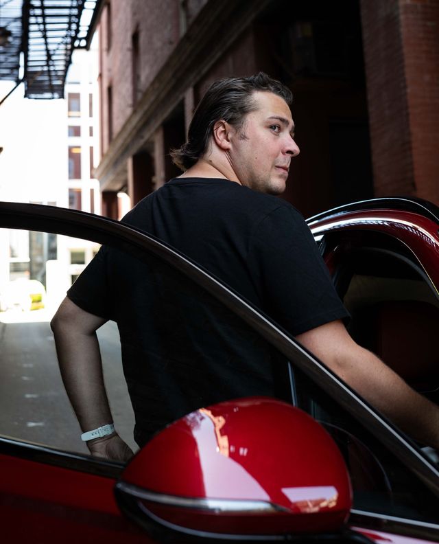 Man in a black shirt standing by a red car in a narrow alley, seen through the open door