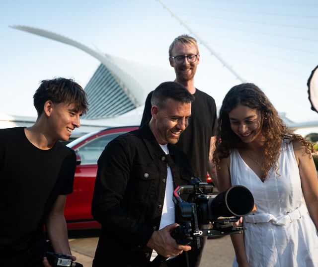 Four people smiling and looking at a camera on a bridge, with a red car and city skyline behind them