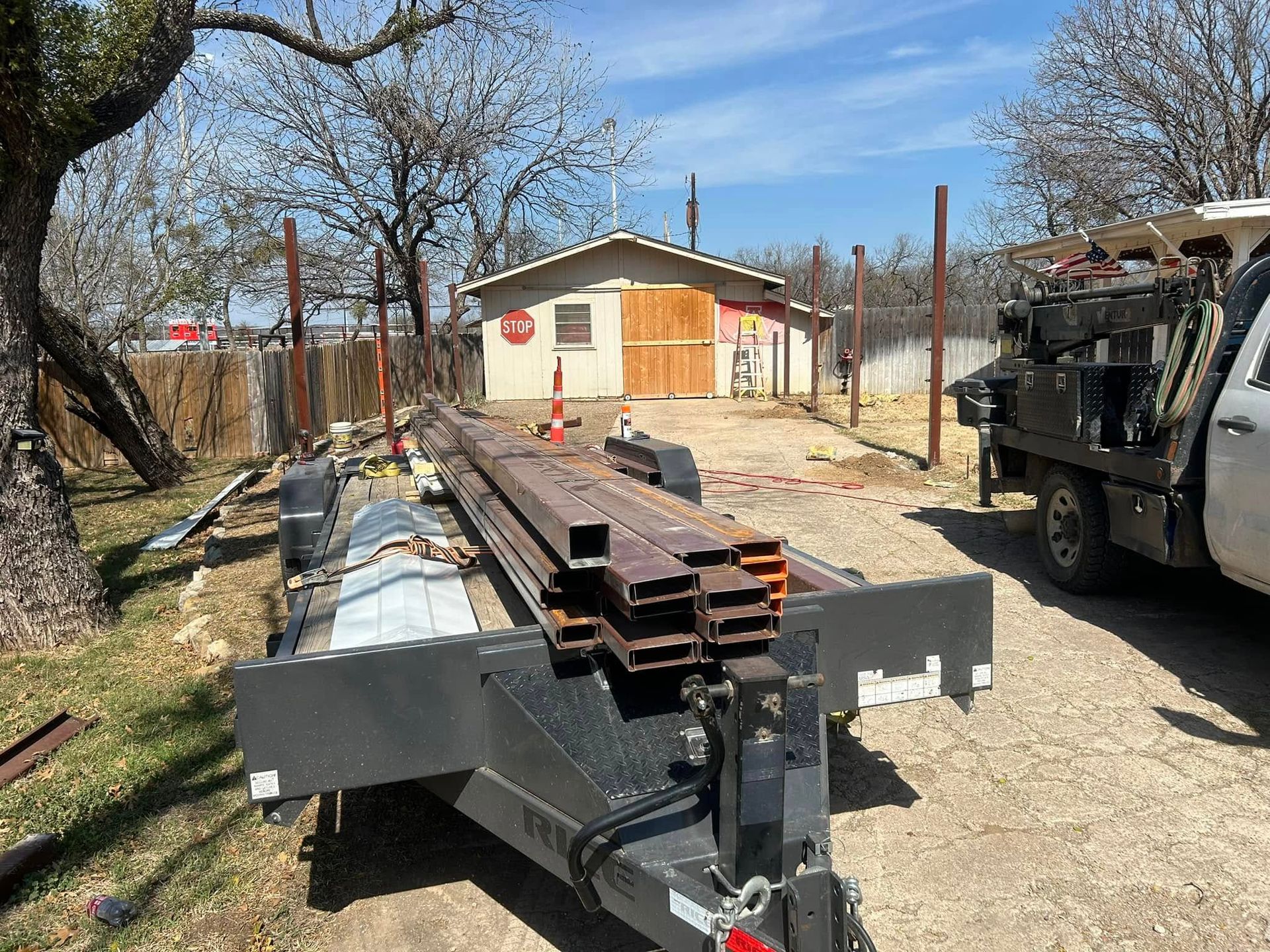 A flatbed trailer loaded with steel beams sits on a gravel drive near a workshop with tan walls and wooden double doors.
