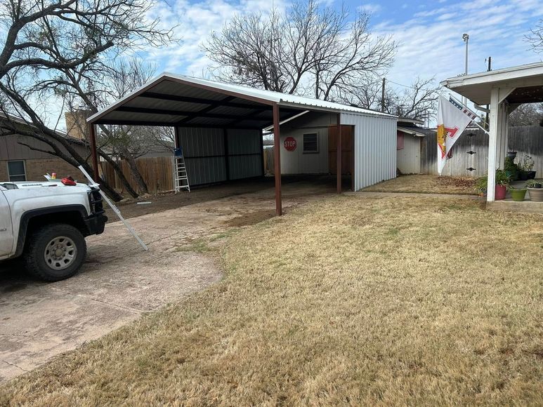 A metal-roofed carport with side panels stands on a dirt and grass lot next to a white truck and a house patio.