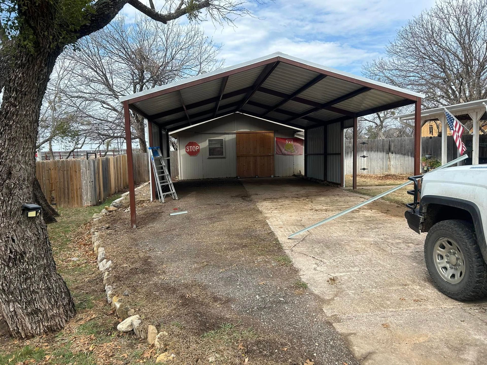 A metal carport structure on a gravel driveway in front of a building, with a ladder leaning against the side.
