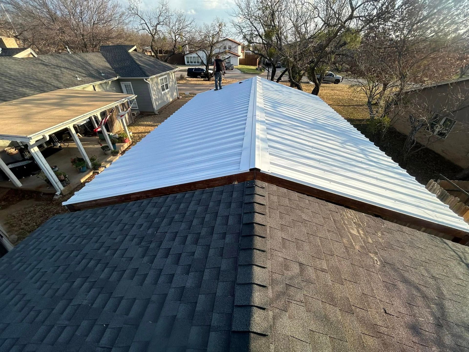 A person stands on a roof partially covered with new white metal roofing panels, transitioning from dark asphalt shingles.