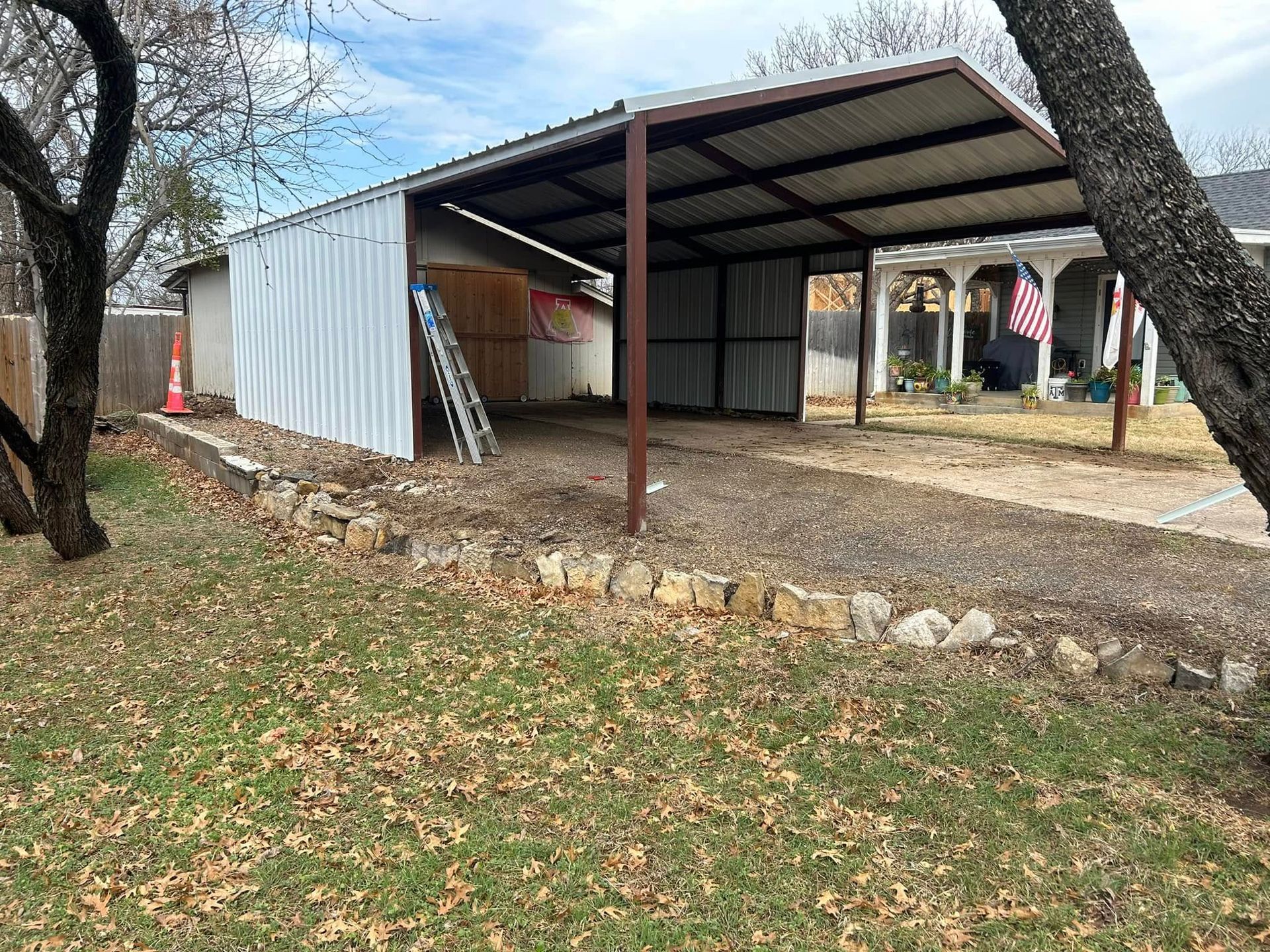 A metal carport with partial siding stands on a gravel driveway in a backyard with trees, a lawn, and a stone border.