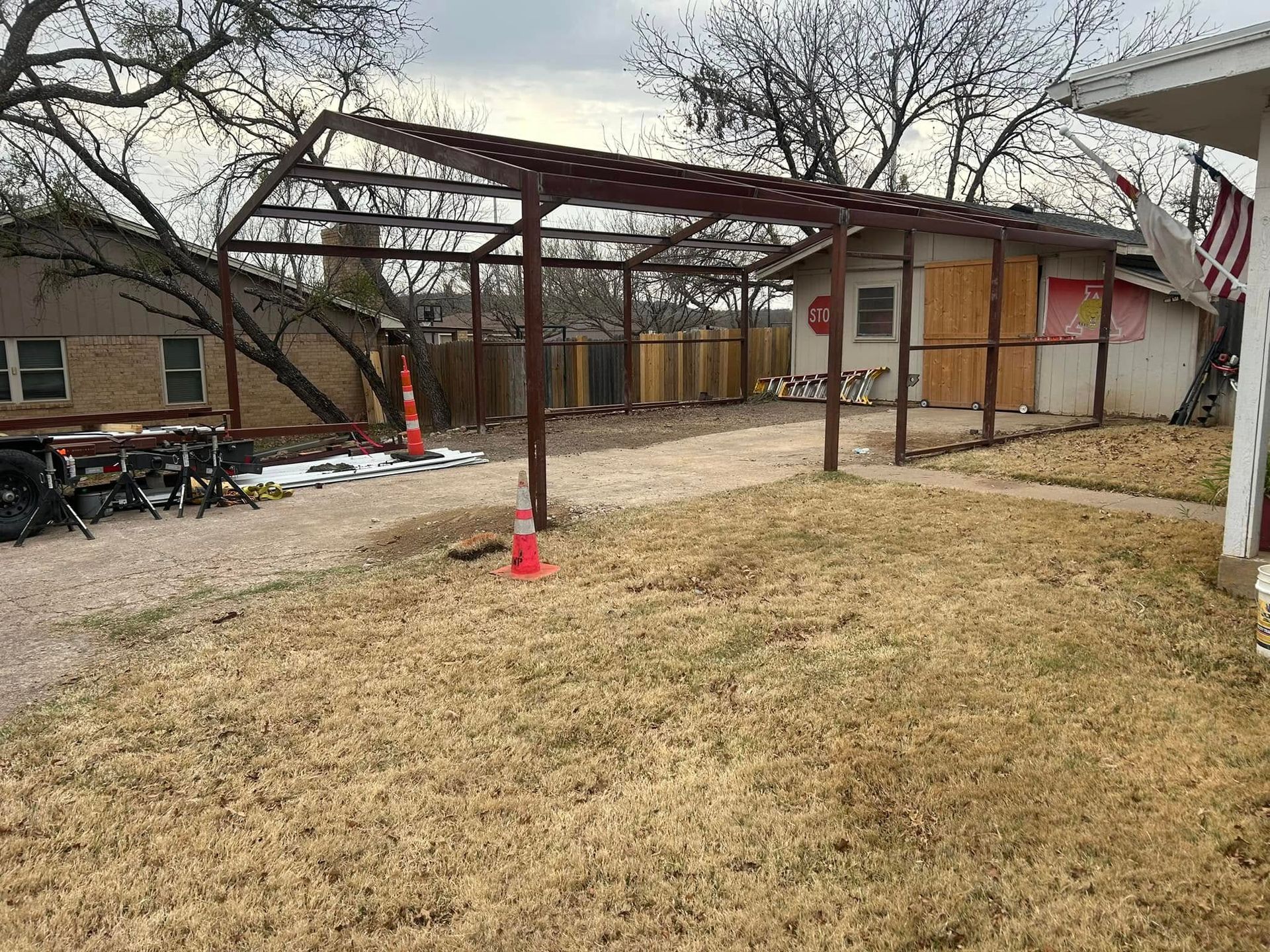 A metal carport frame stands in a backyard next to a house, with orange cones marking the gravel ground.