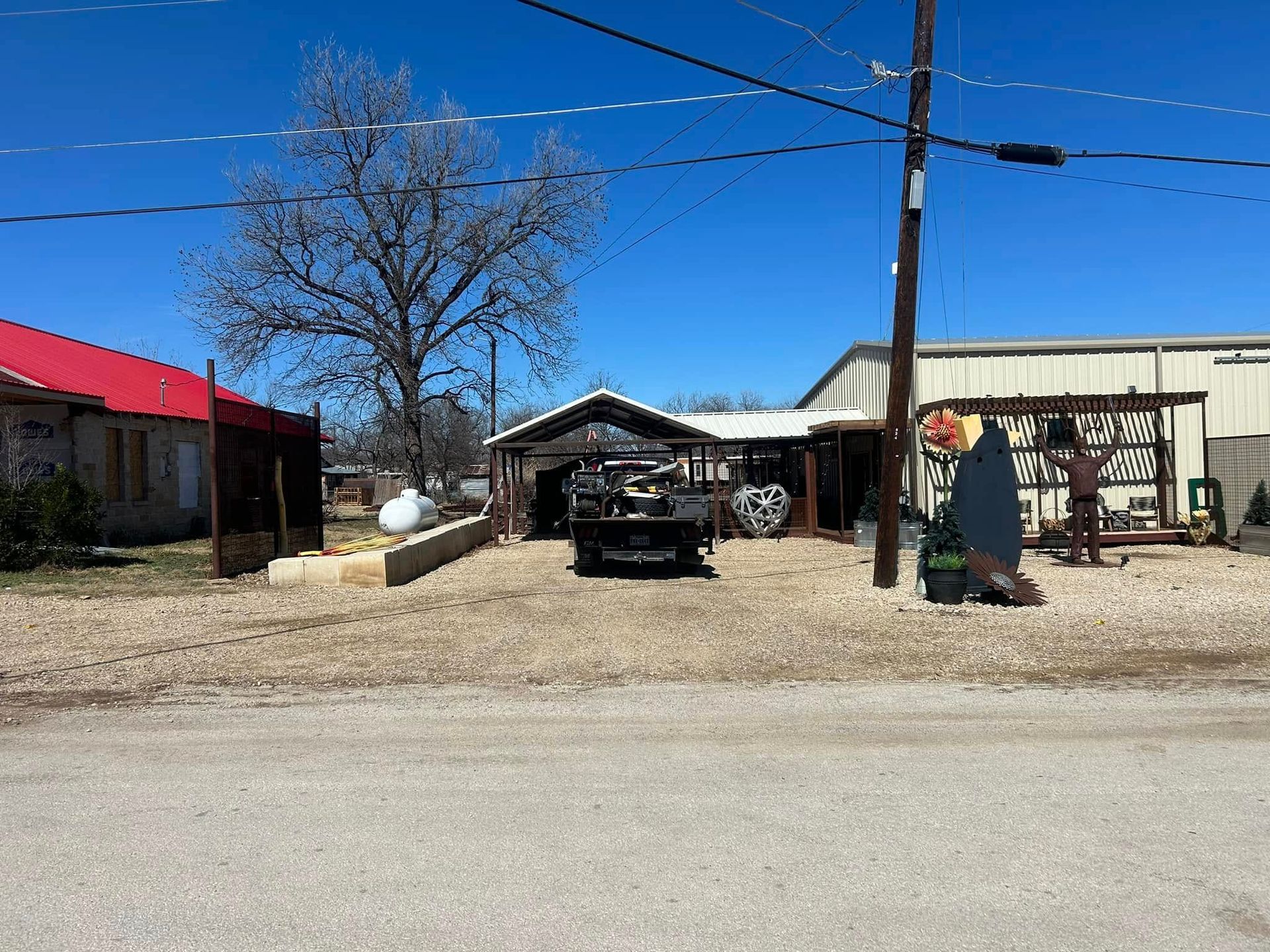 A gravel driveway leads to a carport between a building with a red roof and a metal warehouse on a clear, sunny day.