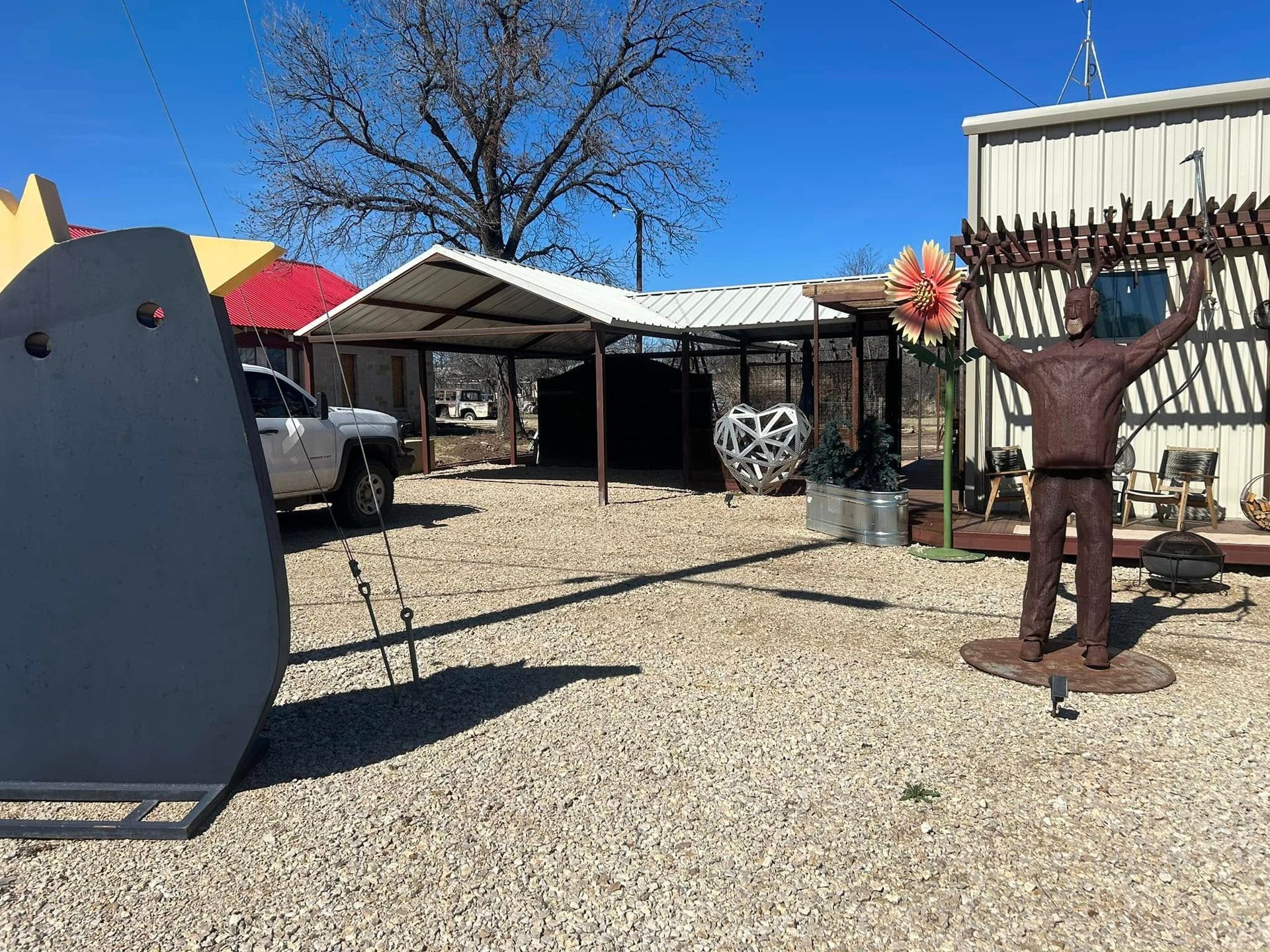 A metal sculpture of a figure with raised arms stands in a gravel lot next to a covered outdoor structure under a blue sky.
